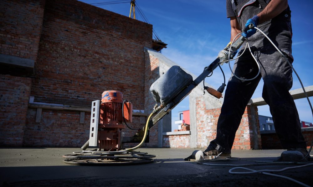 Cropped picture of man laborer in work overalls using power trowel machine while screeding floor in new building. Male worker finishing concrete surface with troweling machine at construction site.
